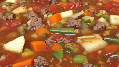 A steaming bowl of ground beef vegetable soup, filled with carrots, potatoes, green beans, and tomatoes, garnished with fresh herbs."