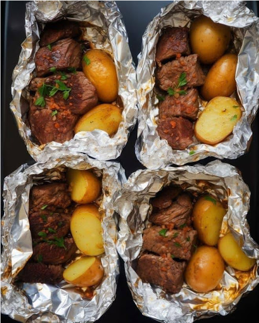 Garlic Steak & Potato Foil Packets fresh off the grill, with steam rising as they're opened. The golden potatoes and juicy steak cubes are visibly seasoned with herbs and spices, ready to be enjoyed in a casual outdoor setting