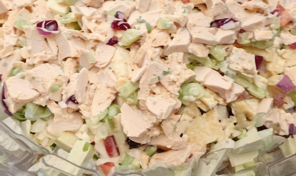 A vibrant and fresh deli-style chicken salad served in a white bowl, garnished with slices of celery and a sprinkle of black pepper, set on a picnic table with a light, airy summer backdrop