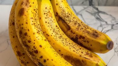 A vibrant, close-up image of a peeled ripe banana with brown spots on its yellow peel, resting on a wooden cutting board alongside a bowl of sliced bananas. The background features ingredients like a glass of almond milk, a handful of nuts, and a drizzle of honey, suggesting versatile ways to incorporate bananas into meals