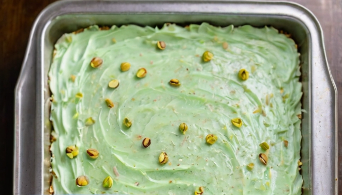 A freshly frosted pineapple pistachio cake sits in a glass baking dish, garnished with chopped pistachios and a light green frosting that glistens under soft lighting. Slices are cut to reveal the moist, airy texture of the cake, with pineapple chunks visible within.