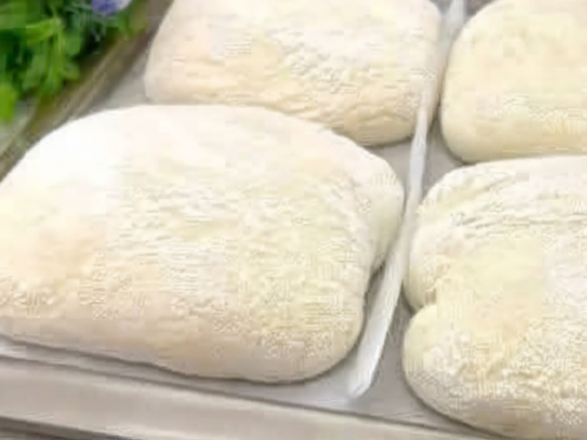 A close-up shot of rustic ciabatta loaves resting on a wooden board, their golden-brown crusts dusted lightly with flour. Sliced open to reveal the airy crumb, with steam rising to suggest freshness.