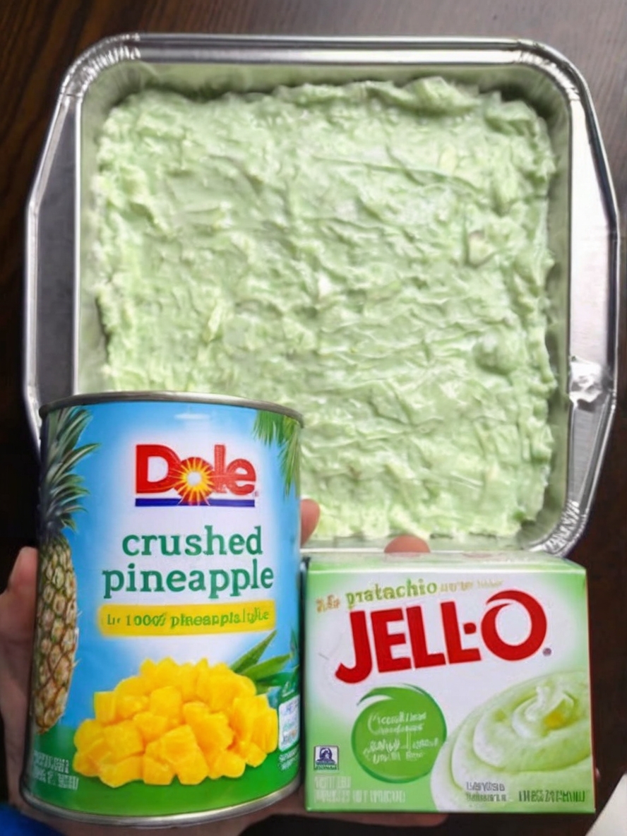A freshly frosted pineapple pistachio cake sits in a glass baking dish, garnished with chopped pistachios and a light green frosting that glistens under soft lighting. Slices are cut to reveal the moist, airy texture of the cake, with pineapple chunks visible within.