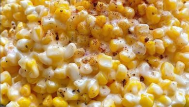 A skillet filled with vibrant, golden corn kernels coated in a glossy honey-butter sauce. Small chunks of cream cheese are melted throughout, adding a creamy texture. The dish is garnished with a sprinkle of freshly cracked black pepper and a sprig of parsley for a fresh, appealing presentation.