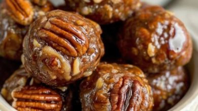 A close-up of golden-brown Pecan Pie Balls coated in powdered sugar, arranged neatly on a rustic wooden plate. A scattering of chopped pecans and a cinnamon stick are styled around the plate, with warm lighting to create a cozy and inviting atmosphere.