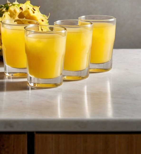 Vibrant Pineapple Ginger Shots lined up on a kitchen counter, each filled with the bright yellow blend. The background shows fresh pineapples, ginger root, and a halved lime, emphasizing the freshness and naturalness of the ingredients.