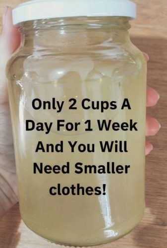 Image is a close-up photograph of a hand holding a clear glass jar filled with a light yellow liquid. The jar has a white plastic lid and is held against a wooden background. The hand visible in the image has light skin. Overlaid on the jar is bold black text in a sans-serif font that reads: "Only 2 Cups A Day For 1 Week And You Will Need Smaller clothes!" The text is centered and takes up most of the visible space on the jar, making it the focal point of the image.