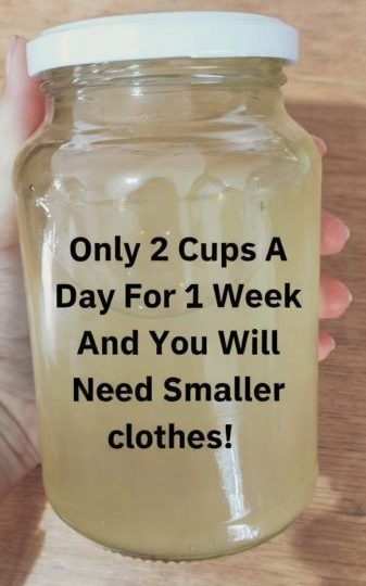 Image is a close-up photograph of a hand holding a clear glass jar filled with a light yellow liquid. The jar has a white plastic lid and is held against a wooden background. The hand visible in the image has light skin. Overlaid on the jar is bold black text in a sans-serif font that reads: "Only 2 Cups A Day For 1 Week And You Will Need Smaller clothes!" The text is centered and takes up most of the visible space on the jar, making it the focal point of the image.