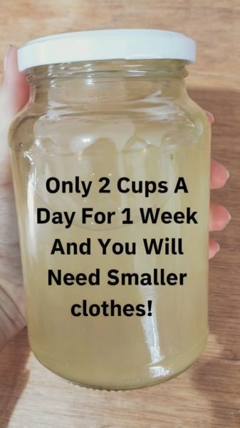 Image is a close-up photograph of a hand holding a clear glass jar filled with a light yellow liquid. The jar has a white plastic lid and is held against a wooden background. The hand visible in the image has light skin. Overlaid on the jar is bold black text in a sans-serif font that reads: "Only 2 Cups A Day For 1 Week And You Will Need Smaller clothes!" The text is centered and takes up most of the visible space on the jar, making it the focal point of the image.