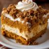 Pumpkin cream cheese crumble cake with spiced pumpkin cake, creamy cheesecake-style filling, golden cinnamon crumble topping, and whipped cream dusted with cinnamon.