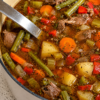 A steaming bowl of hearty vegetable beef soup filled with tender chunks of seared beef, diced carrots, potatoes, green beans, and corn in a rich savory broth, garnished with fresh chopped parsley.