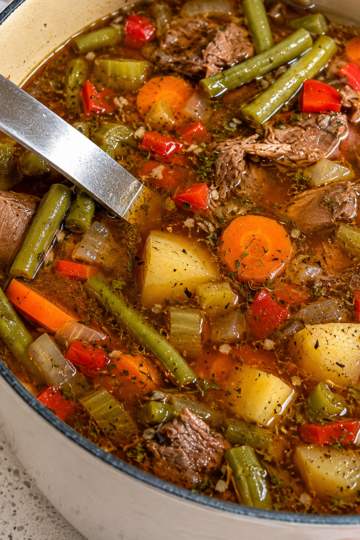 A steaming bowl of hearty vegetable beef soup filled with tender chunks of seared beef, diced carrots, potatoes, green beans, and corn in a rich savory broth, garnished with fresh chopped parsley.