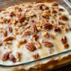 "Butter pecan poke cake topped with creamy vanilla glaze and generously sprinkled with chopped pecans, served in a glass baking dish for a classic Southern homemade dessert."