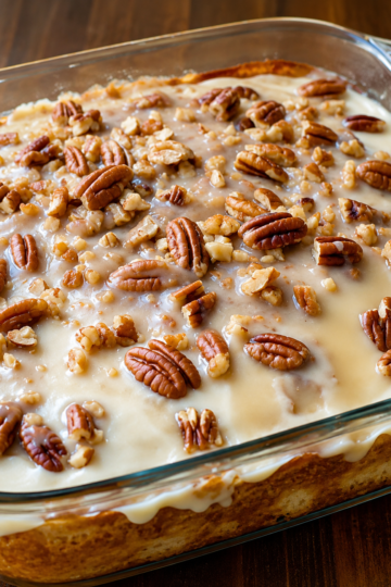 "Butter pecan poke cake topped with creamy vanilla glaze and generously sprinkled with chopped pecans, served in a glass baking dish for a classic Southern homemade dessert."