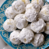 "Powdered sugar snowball cookies piled on a blue decorative plate, featuring buttery homemade cookies generously coated in confectioners' sugar."