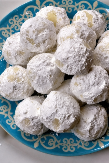 "Powdered sugar snowball cookies piled on a blue decorative plate, featuring buttery homemade cookies generously coated in confectioners' sugar."