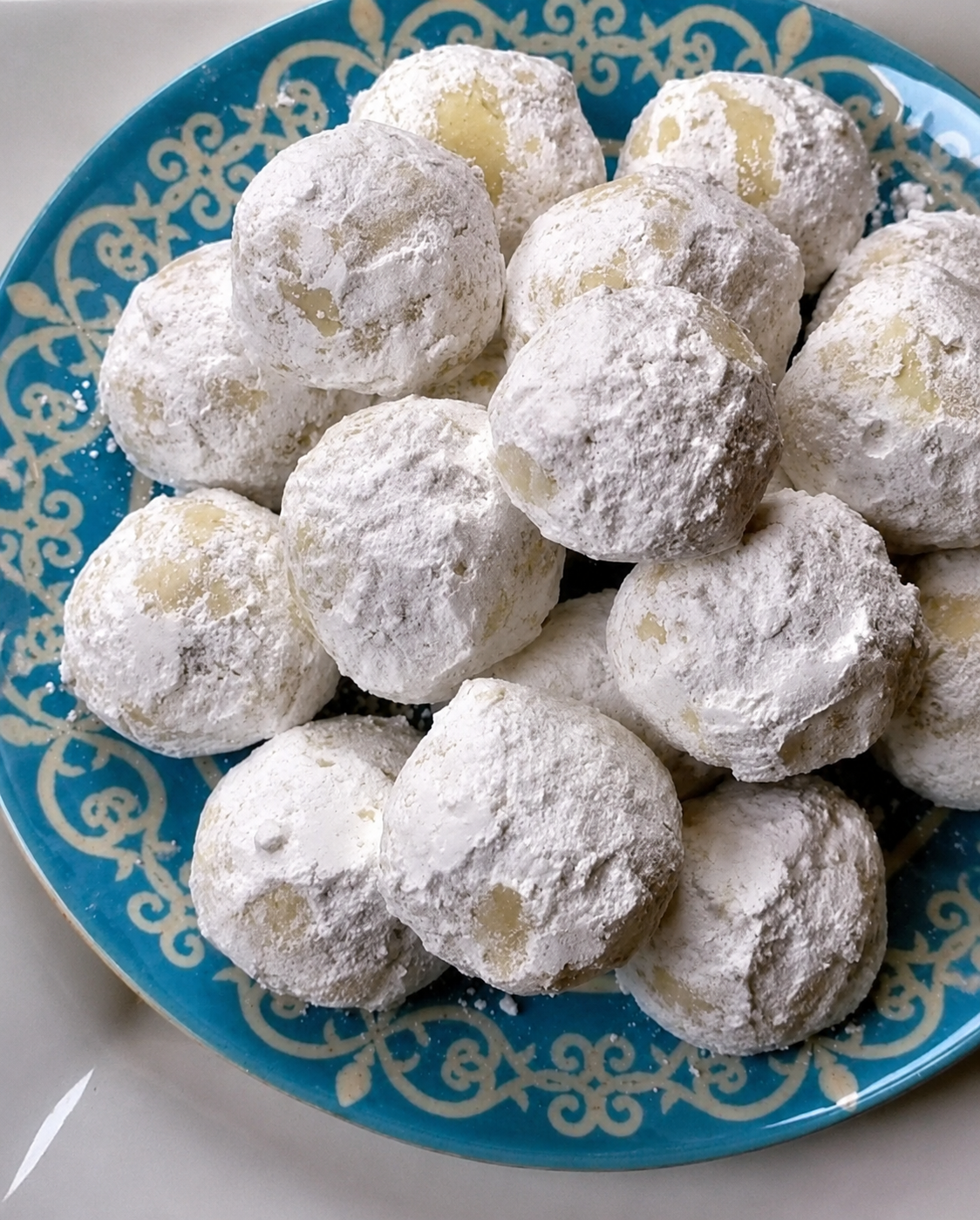 "Powdered sugar snowball cookies piled on a blue decorative plate, featuring buttery homemade cookies generously coated in confectioners' sugar."