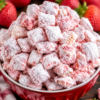 Close-up of a bowl filled with Strawberry Shortcake Puppy Chow, featuring crunchy Chex cereal coated in a sweet strawberry-flavored white chocolate mixture and generously dusted with powdered sugar. Fresh strawberries are softly blurred in the background, highlighting the dessert's vibrant pink and white colors.