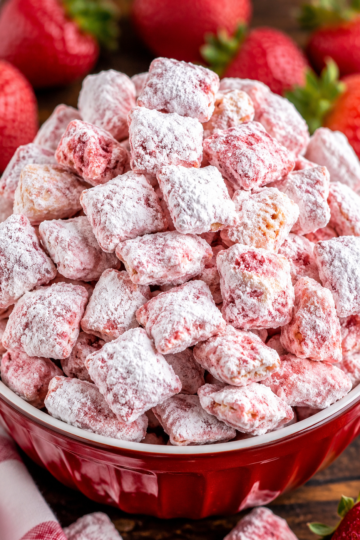 Close-up of a bowl filled with Strawberry Shortcake Puppy Chow, featuring crunchy Chex cereal coated in a sweet strawberry-flavored white chocolate mixture and generously dusted with powdered sugar. Fresh strawberries are softly blurred in the background, highlighting the dessert's vibrant pink and white colors.