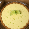 Slice of classic key lime pie with a golden graham cracker crust, creamy pale-yellow lime filling, topped with thin fresh lime slices and a sprinkle of lime zest on a white plate.