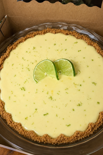Slice of classic key lime pie with a golden graham cracker crust, creamy pale-yellow lime filling, topped with thin fresh lime slices and a sprinkle of lime zest on a white plate.