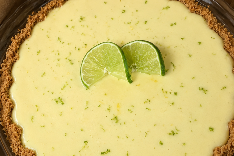 Slice of classic key lime pie with a golden graham cracker crust, creamy pale-yellow lime filling, topped with thin fresh lime slices and a sprinkle of lime zest on a white plate.