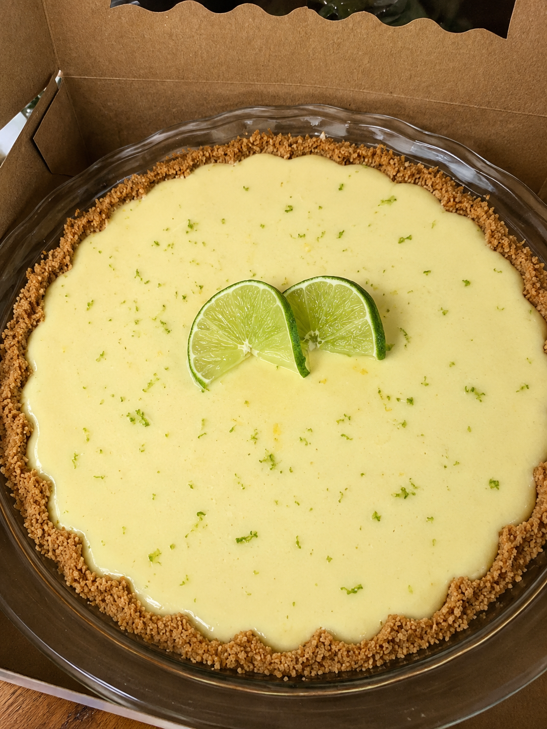 Slice of classic key lime pie with a golden graham cracker crust, creamy pale-yellow lime filling, topped with thin fresh lime slices and a sprinkle of lime zest on a white plate.
