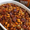 Homemade baked beans with crispy bacon and caramelized onions in a rich sweet and smoky sauce, served in a white baking dish for a classic Southern comfort food side dish.
