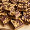 The image has been recreated with a realistic homemade look, showing rich chocolate oat bars stacked on a wooden board with glossy chocolate and chewy oat layers.