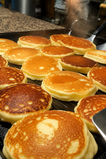 A tall stack of fluffy sourdough pancakes with crispy golden edges, topped with melting butter and drizzled with pure maple syrup. Served with fresh berries and a dusting of powdered sugar on a rustic plate, photographed in warm morning natural light with a cozy farmhouse breakfast setting.