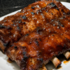 Tender Crockpot Barbecue Ribs coated in a glossy homemade barbecue sauce, slow-cooked until fall-off-the-bone and finished under the broiler for a sticky caramelized glaze. Served on a rustic wooden platter with extra barbecue sauce, fresh parsley, and classic sides like coleslaw and cornbread. Photographed in warm natural light, highlighting the juicy meat, rich smoky sauce, and irresistible barbecue finish.