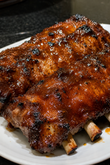Tender Crockpot Barbecue Ribs coated in a glossy homemade barbecue sauce, slow-cooked until fall-off-the-bone and finished under the broiler for a sticky caramelized glaze. Served on a rustic wooden platter with extra barbecue sauce, fresh parsley, and classic sides like coleslaw and cornbread. Photographed in warm natural light, highlighting the juicy meat, rich smoky sauce, and irresistible barbecue finish.