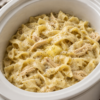 A cozy bowl of Slow Cooker Amish Chicken and Noodles featuring tender shredded chicken and thick homestyle egg noodles in a rich, creamy broth. Garnished with freshly chopped parsley and served with warm buttered biscuits on a rustic wooden table in soft natural light, creating the perfect comforting homemade family dinner.