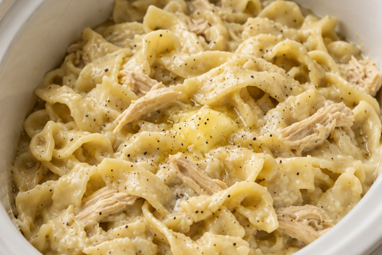 A cozy bowl of Slow Cooker Amish Chicken and Noodles featuring tender shredded chicken and thick homestyle egg noodles in a rich, creamy broth. Garnished with freshly chopped parsley and served with warm buttered biscuits on a rustic wooden table in soft natural light, creating the perfect comforting homemade family dinner.