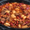 A hearty bowl of Crockpot Calico Beans made with kidney beans, cannellini beans, pork and beans, crispy bacon, and optional ground beef in a thick, glossy sweet-and-tangy sauce. Garnished with chopped parsley and served with a slice of warm cornbread on a rustic wooden table in cozy natural light, highlighting the rich, smoky flavors of this classic slow-cooker comfort food