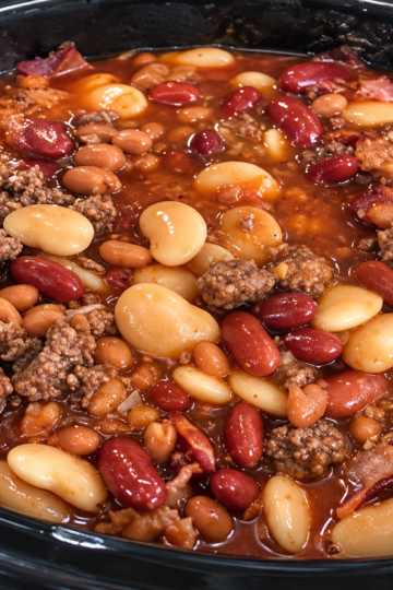 A hearty bowl of Crockpot Calico Beans made with kidney beans, cannellini beans, pork and beans, crispy bacon, and optional ground beef in a thick, glossy sweet-and-tangy sauce. Garnished with chopped parsley and served with a slice of warm cornbread on a rustic wooden table in cozy natural light, highlighting the rich, smoky flavors of this classic slow-cooker comfort food