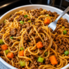 A close-up of homemade ground beef lo mein served in a rustic bowl, featuring tender spaghetti noodles coated in a glossy soy-garlic sauce with seasoned ground beef, crisp carrots, celery, and red onions. Garnished with sliced green onions and sesame seeds, the dish is photographed under warm natural light on a wooden table for a cozy, better-than-takeout presentation.