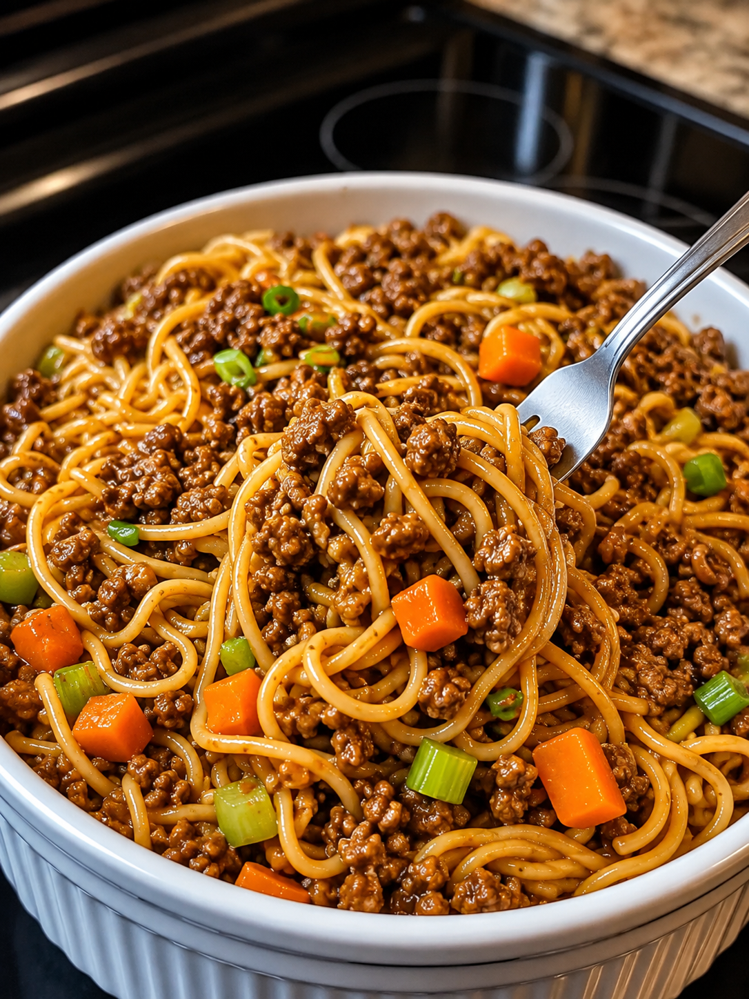 A close-up of homemade ground beef lo mein served in a rustic bowl, featuring tender spaghetti noodles coated in a glossy soy-garlic sauce with seasoned ground beef, crisp carrots, celery, and red onions. Garnished with sliced green onions and sesame seeds, the dish is photographed under warm natural light on a wooden table for a cozy, better-than-takeout presentation.