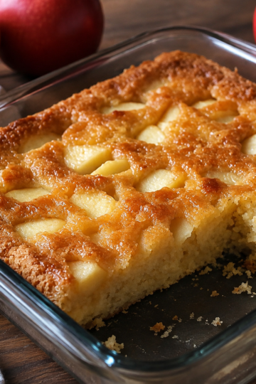 A moist homemade apple cinnamon cake filled with tender chunks of fresh apple and topped with a light dusting of powdered sugar. One slice is cut to reveal the soft, buttery crumb with juicy apple pieces and optional chopped walnuts throughout. Served on a rustic wooden cake stand with fresh apple slices and cinnamon sticks, photographed in warm natural light for a cozy fall dessert presentation.