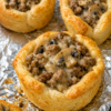 Golden, flaky crescent roll cups baked in a muffin tin and filled with a creamy mixture of seasoned ground beef, sautéed mushrooms, cream cheese, and melted cheddar cheese. One crescent cup is cut open to reveal the rich, savory filling, while the others are topped with bubbling golden cheese and fresh parsley. Served on a rustic wooden board in warm natural light, creating a cozy homemade appetizer or weeknight dinner presentation.