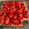 A slice of homemade strawberry graham cracker icebox cake showing soft, cake-like layers of graham crackers, fluffy vanilla whipped cream, and fresh sliced strawberries. The no-bake dessert is topped with extra strawberries and served chilled on a white dessert plate, photographed in warm natural light on a rustic wooden table for a fresh, summery homemade presentation.