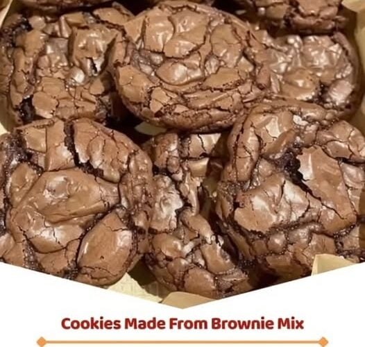 A stack of fudgy brownie mix cookies with shiny, crackly tops, soft chocolate centers, and slightly crisp edges. One cookie is broken in half to reveal the rich, gooey interior with melted chocolate throughout. Arranged on a cooling rack with scattered chocolate chips on a rustic wooden table, photographed in warm natural light for a cozy homemade dessert presentation.
