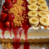 A no-bake split cheesecake in a 9x13-inch pan featuring two delicious halves: one topped with fresh strawberries, glossy strawberry glaze, and crunchy golden cookie crumbs, and the other layered with sliced bananas and vanilla wafer crumbs. The creamy cheesecake filling rests on a buttery vanilla wafer crust, with neatly cut squares revealing the smooth, mousse-like texture. Served chilled and photographed in warm natural light for a bakery-style dessert presentation.