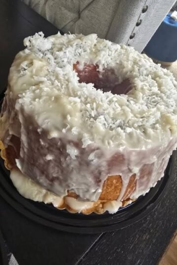 A moist coconut rum pound cake coated in creamy white glaze and generously covered with shredded coconut. The golden cake is served whole with a large slice missing, showing its rich, tender interior.