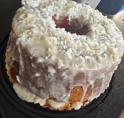 A moist coconut rum pound cake coated in creamy white glaze and generously covered with shredded coconut. The golden cake is served whole with a large slice missing, showing its rich, tender interior.