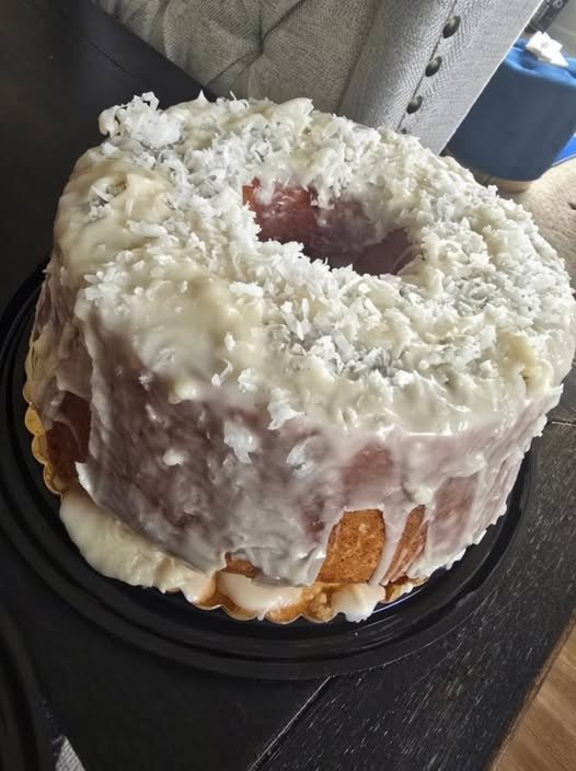 A moist coconut rum pound cake coated in creamy white glaze and generously covered with shredded coconut. The golden cake is served whole with a large slice missing, showing its rich, tender interior.