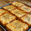Close-up of cheesy garlic Texas toast made with thick slices of buttery bread, topped with melted mozzarella, provolone, and Parmesan cheese. The toast is baked until golden and bubbly with crispy edges, then sprinkled with dried parsley and served on a rustic wooden board in warm natural light. Long, gooey cheese pulls and a rich garlic butter topping create an irresistible homemade side dish.
