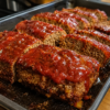 A rustic sheet pan meatloaf glazed with ketchup and baked until deeply caramelized around the edges. Thick slices reveal a juicy, tender interior made with seasoned ground beef, served alongside creamy mashed potatoes and green beans. Garnished with fresh parsley and photographed in warm natural light on a wooden farmhouse table, creating a classic homemade comfort food presentation.