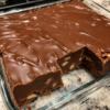 Close-up of rich old-fashioned chocolate fudge cut into neat squares and arranged on a rustic serving plate. The creamy homemade fudge is loaded with chopped nuts and has a smooth, glossy chocolate finish. Photographed in warm natural light with a few extra fudge pieces and chocolate chips scattered on a wooden table, creating a cozy holiday dessert presentation.