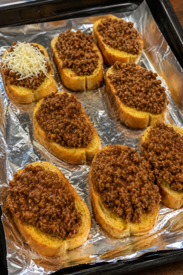 Open-faced Texas Toast Sloppy Joes made with thick slices of buttery golden Texas toast topped with a rich, savory ground beef and onion mixture in a tangy tomato sauce. Finished with melted shredded cheddar cheese and served on a rustic wooden board with pickles and crispy fries. Photographed in warm natural light, showcasing the crisp toast, juicy beef, and gooey cheese for a comforting homemade family dinner.